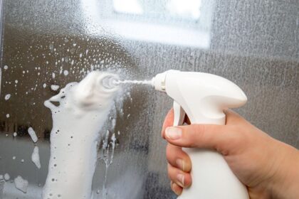 Egy kéz spray-t használ, hogy vízkőoldót fújjon egy üvegfelületre.