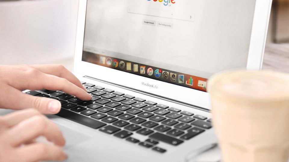 Egy kéz gépel a MacBook Air billentyűzetén, miközben a Google keresőoldala látható a képernyőn.