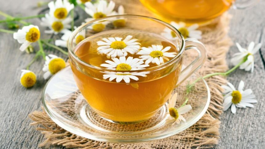 Egy átlátszó pohárban kamilla tea, virágokkal díszítve, fából készült asztalon.