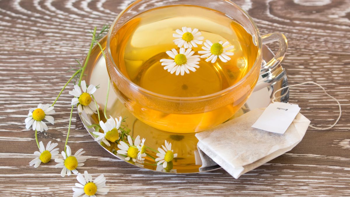 Egy átlátszó bögrében kamillatea látható, virágokkal díszítve, mellette tea filter.