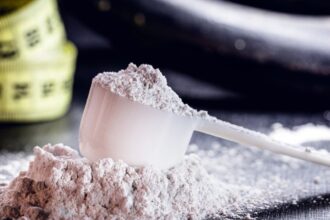 Egy fehér mérőkanál por áll egy halom L-karnitin mellett, háttérben mérőszalag látható.