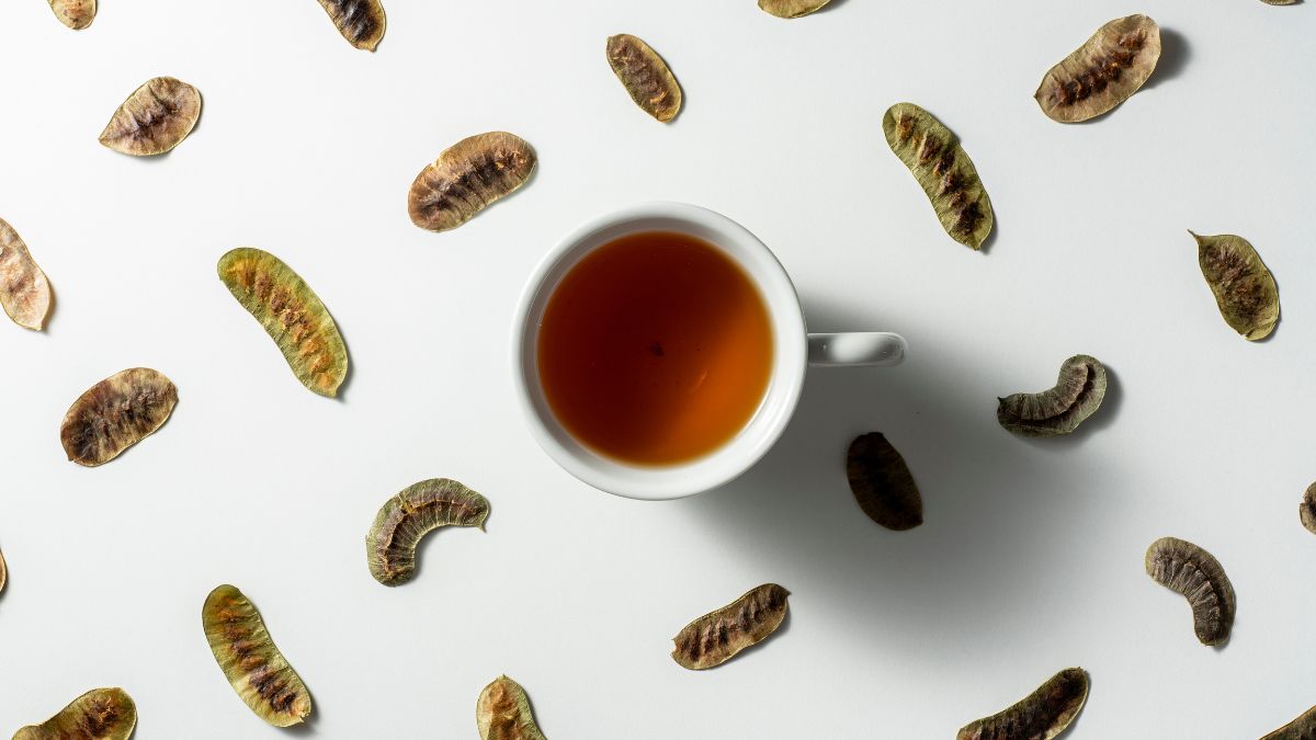 Egy csésze szenna tea, körülötte szenna levelek, világos háttéren.