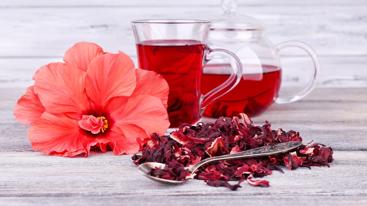 Egy pohár hibiszkusz tea, szárított hibiszkusz virágok és egy virág a háttérben.