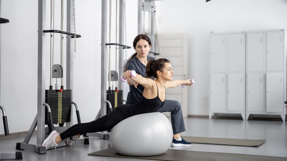 Két nő edz egy fitneszteremben, az egyik nő egy labdán ülve súlyzózik, míg a másik segít neki.