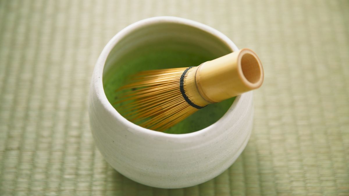 Egy fehér kerámia tálban zöld matcha tea és bambusz habverő látható.
