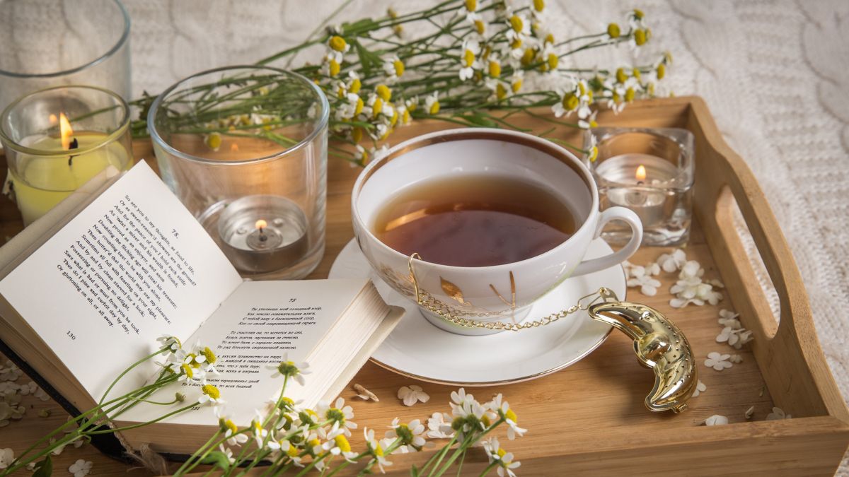 Egy csésze tea, könyv és gyertyák egy fából készült tálcán, virágokkal díszítve.