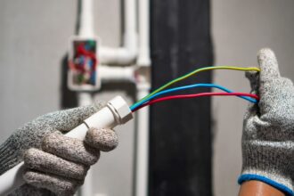 Két kéz védőkesztyűben, elektromos vezetékeket tartva, biztonságos szereléshez.