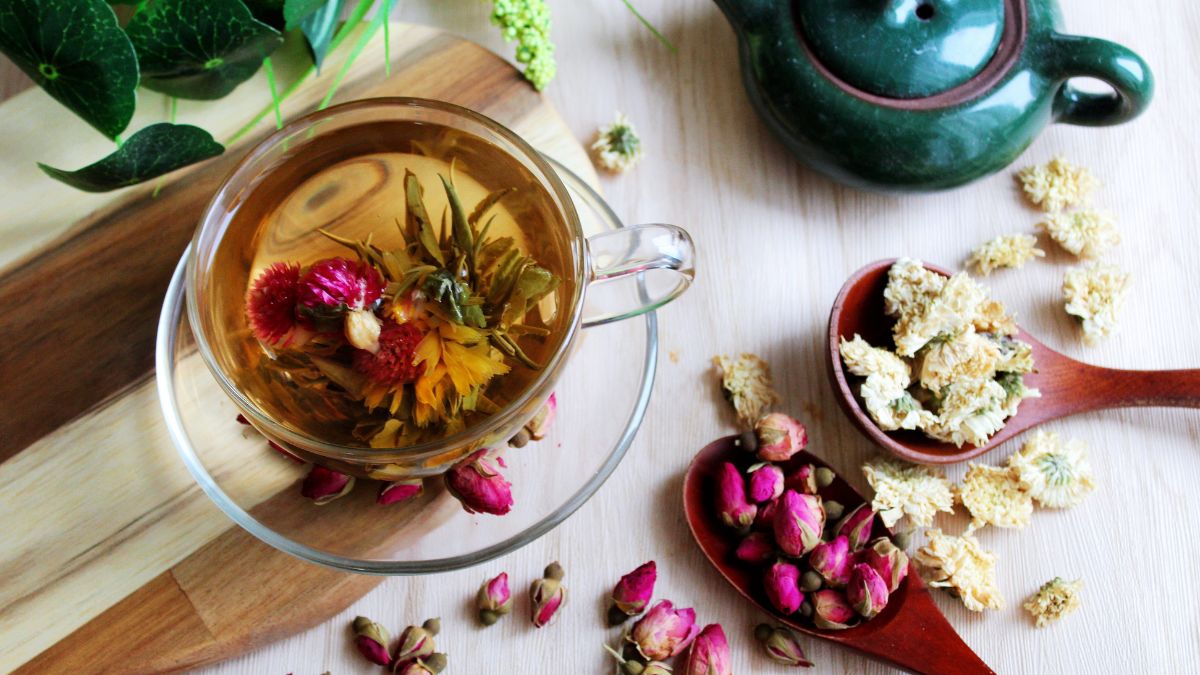 Egy átlátszó pohárban virágos tea, mellette szárított virágok és egy zöld teáskanna.