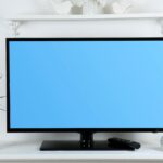 Egy plazma TV látható, kék háttérrel, mellette növény és dísztárgyak.