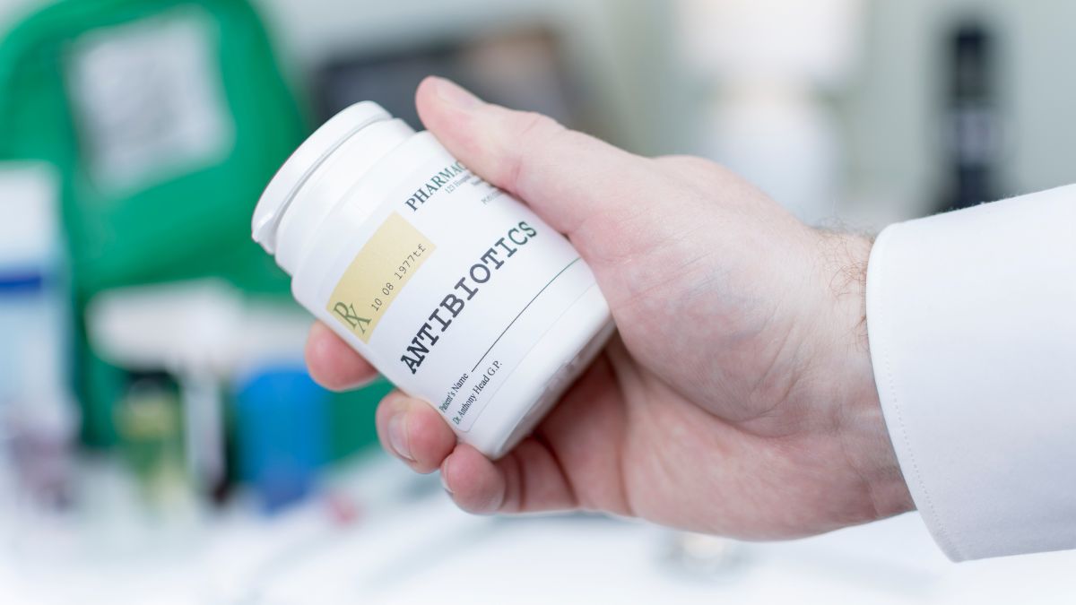 Egy kéz antibiotikumos üveget tart, a háttérben gyógyszerek láthatók.