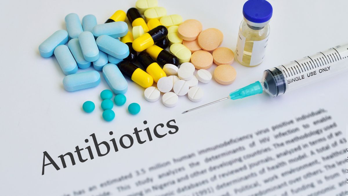 Különböző színű és formájú antibiotikum tabletták, injekciós tű és üveg a gyógyszeres papíron.