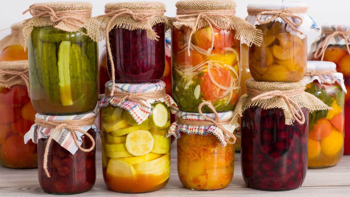 Színes befőtt üvegek sorakoznak, különböző zöldségekkel és gyümölcsökkel, természetes anyagokkal díszítve.