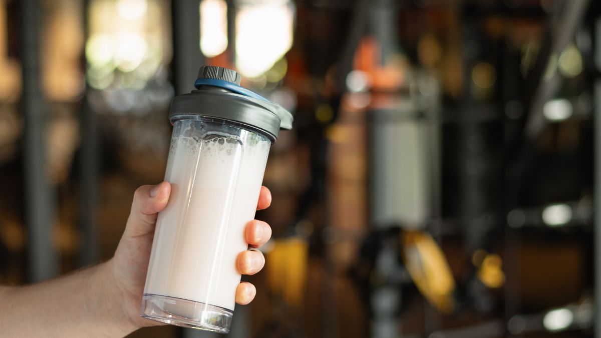 Egy kézben tartott fehér színű fehérje shake shaker, háttérben edzőterem.