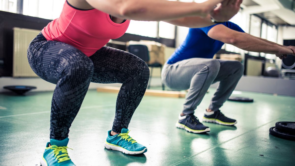 Két ember guggolás közben edzőteremben, különböző sportfelszerelésben.