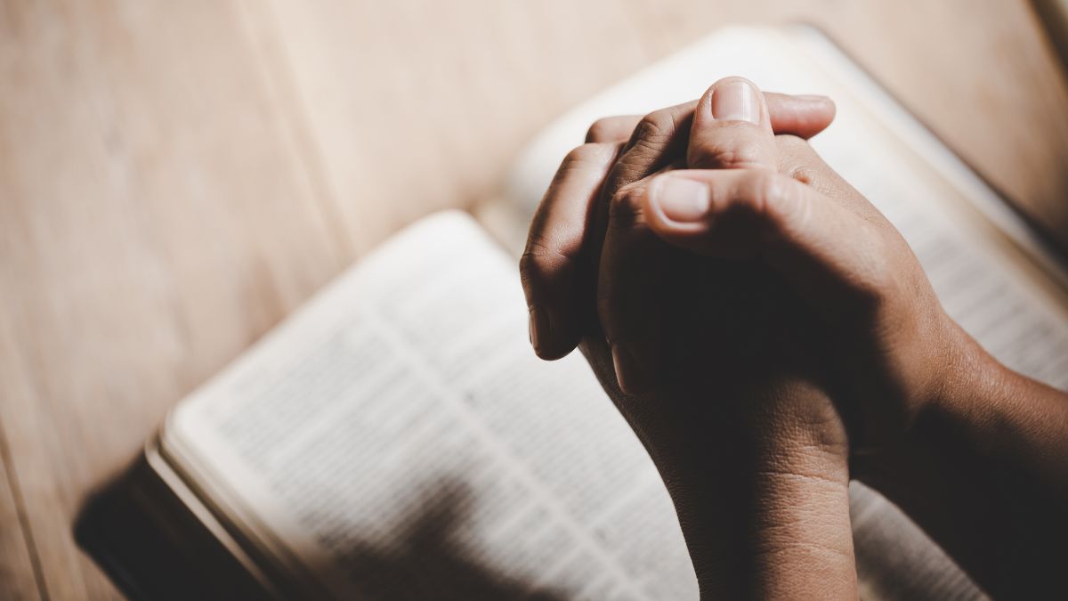 Két kéz összekulcsolva imádkozás közben, egy nyitott Biblia mellett.