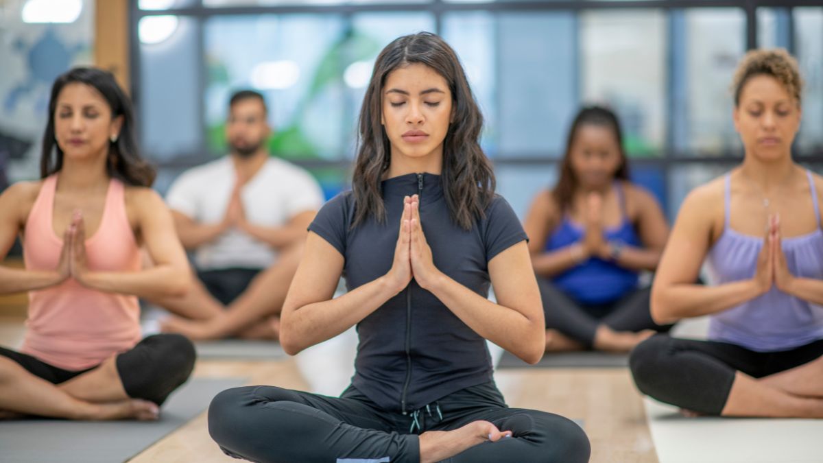 Négy ember jógázik egy stúdióban, meditáló pózban ülve, összekulcsolt kezekkel.