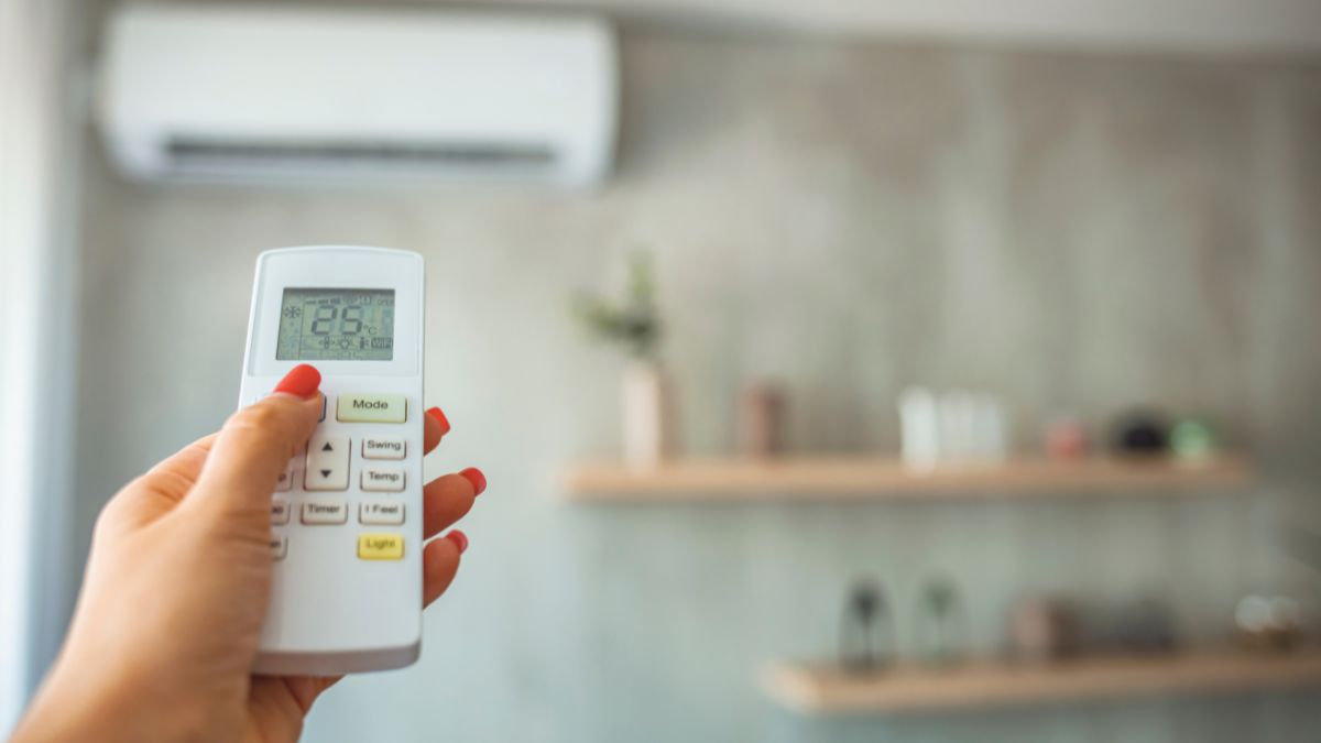 Kézben tartott távirányító, amelyen 25 °C látható, háttérben klímaberendezés.