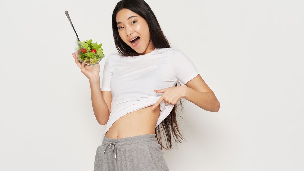 Egy fiatal nő boldogan mutatja a hasát, miközben salátát tart a kezében.
