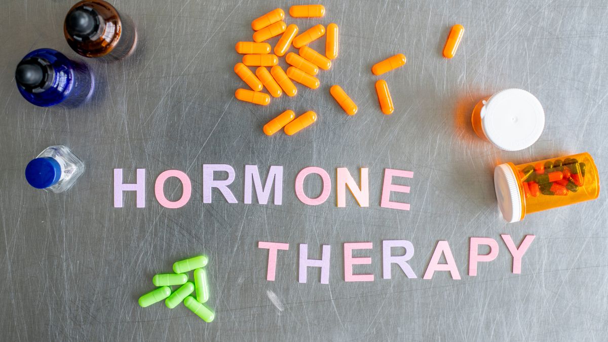 Különböző színű kapszulák és gyógyszerek elhelyezve egy fémfelületen, a 'HORMONE THERAPY' felirattal.