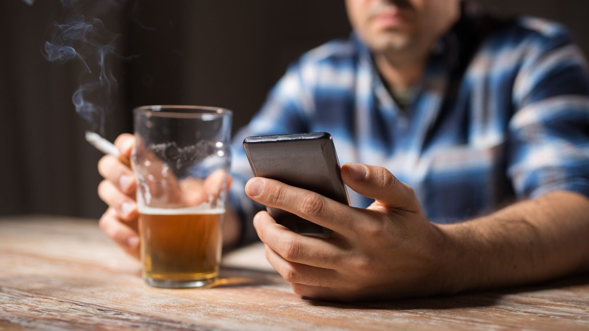 Egy férfi sört tart a kezében, miközben telefonját nézi, füstölve.