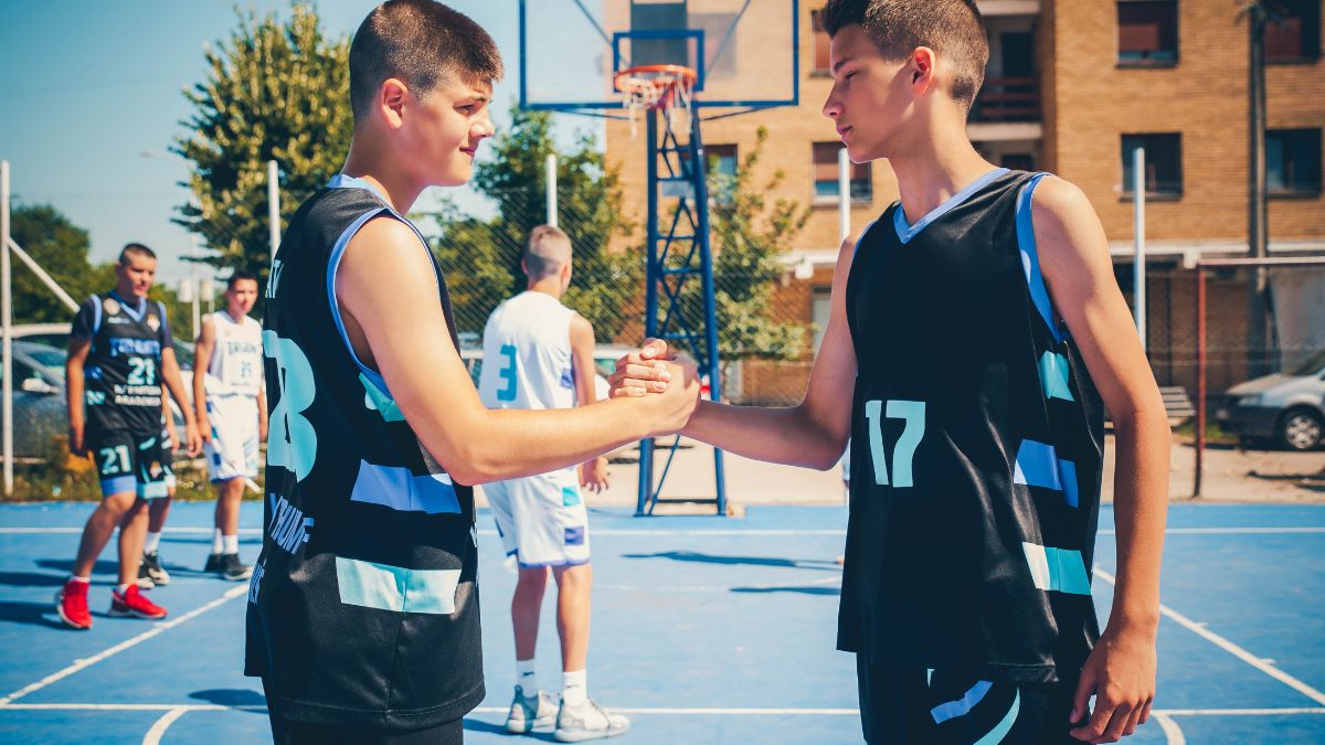 Két fiatal kosárlabdázó kezet fog, miközben a háttérben csapattársaik edzenek.