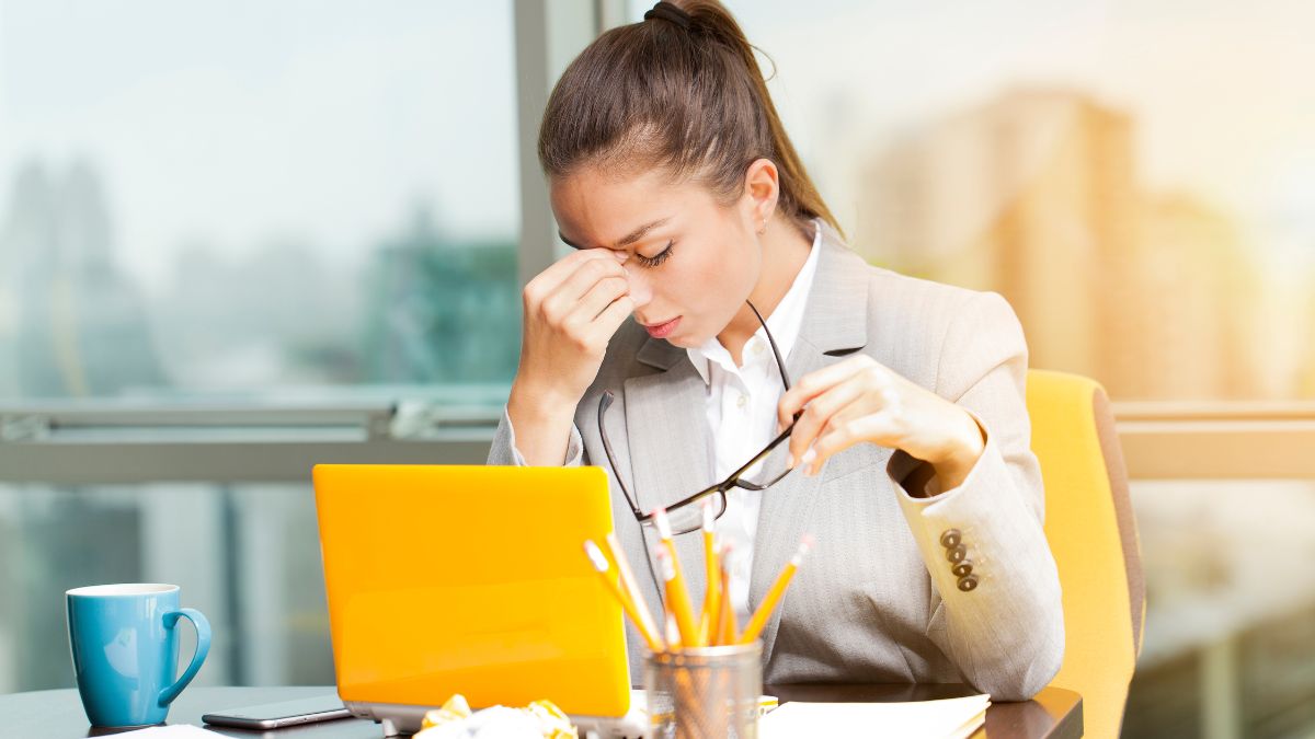 Egy stresszes nő ül az íróasztalánál, kezével a homlokát masszírozza, miközben laptop előtt dolgozik.