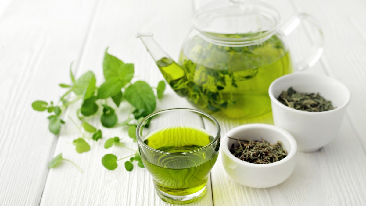 Friss zöld tea egy pohárban, mellette teáskanna és gyógynövények.