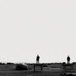 Három álló figura egy üres tájon, körülöttük tárgyakkal.