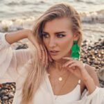 Fiatal nő zöld fülbevalóval áll a vízparton, elegáns stílusban.
